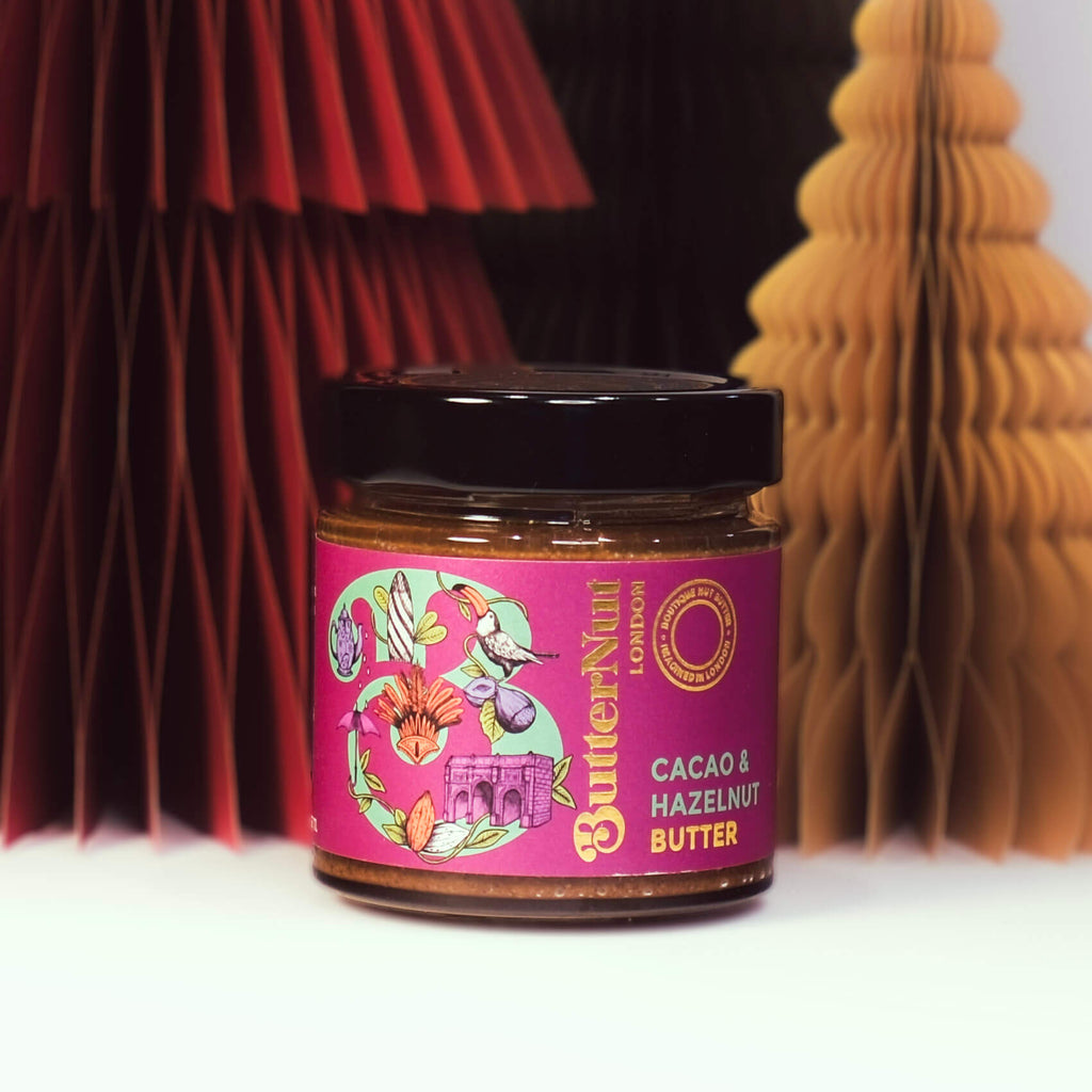 Red, green, and golden paper trees with a jar of ButterNut of London Cacao & Hazelnut Butter on a white background.