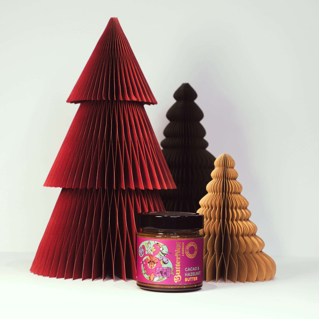 Red, green, and golden paper trees with a jar of ButterNut of London Cacao & Hazelnut Butter on a white background.