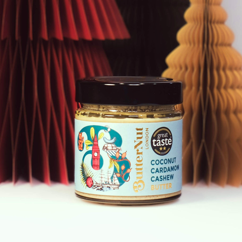 Red, green, and golden paper trees with a jar of ButterNut of London Coconut Cardamom Cashew Butter on a white background.