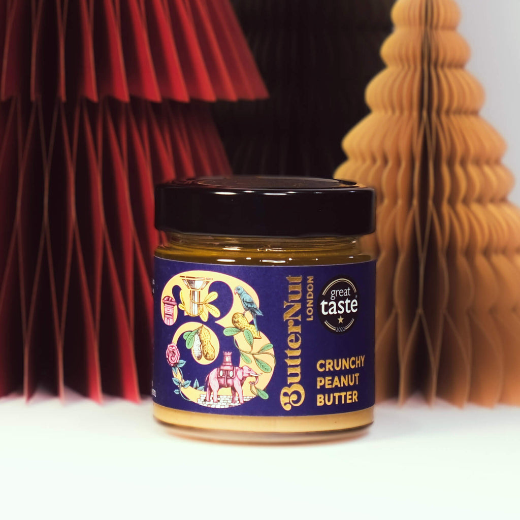 Red, green, and golden paper trees with a jar of ButterNut of London Crunchy Peanut Butter on a white background.