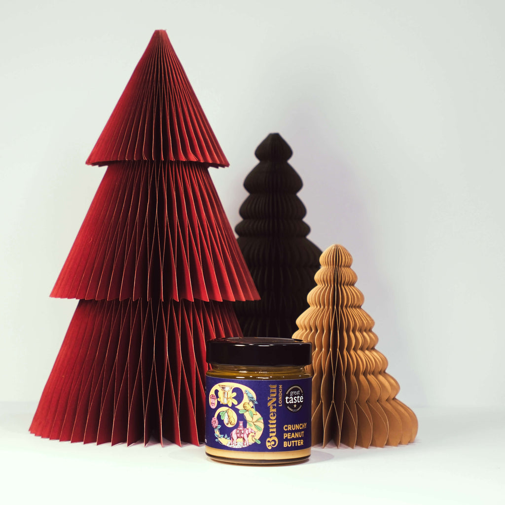 Red, green, and golden paper trees with a jar of ButterNut of London Crunchy Peanut Butter on a white background.