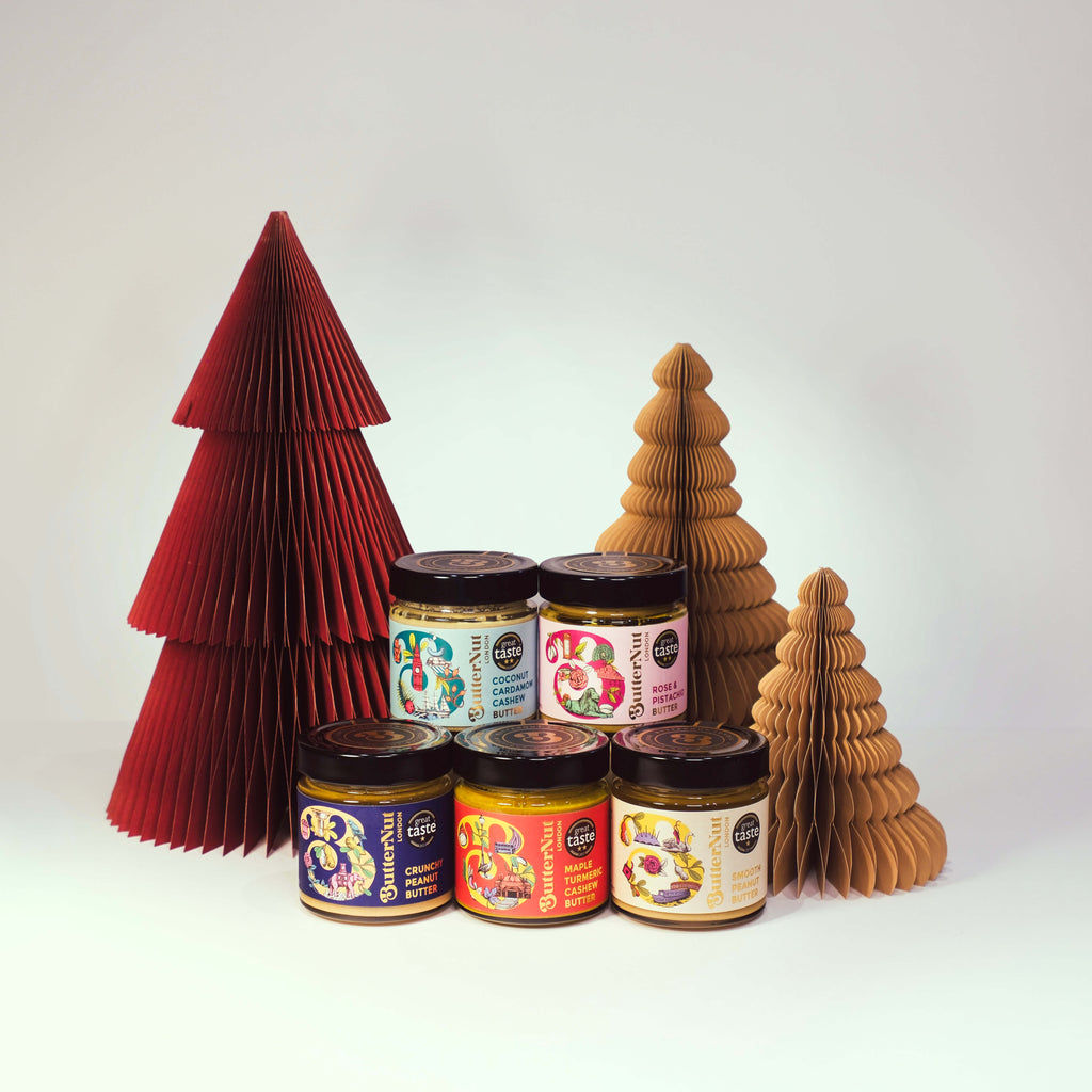Red and golden paper trees with jars of ButterNut of London Great Taste Winning Nut Butters on a white background.