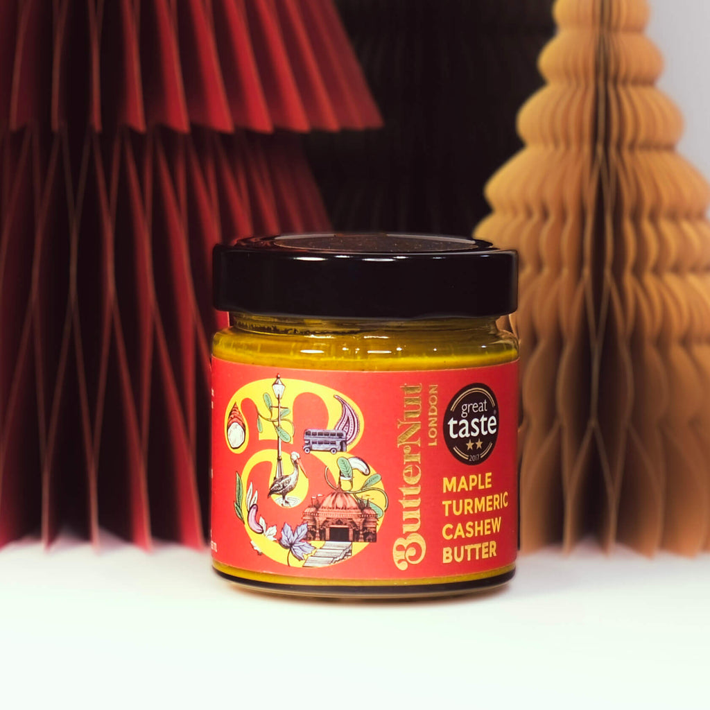 Red, green, and golden paper trees with a jar of ButterNut of London Maple Turmeric Cashew Butter on a white background.