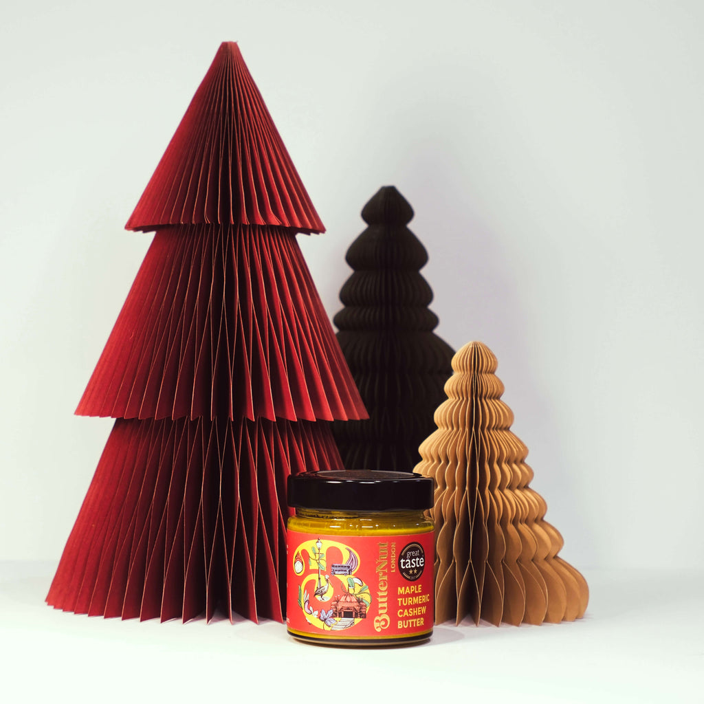 Red, green, and golden paper trees with a jar of ButterNut of London Maple Turmeric Cashew Butter on a white background.