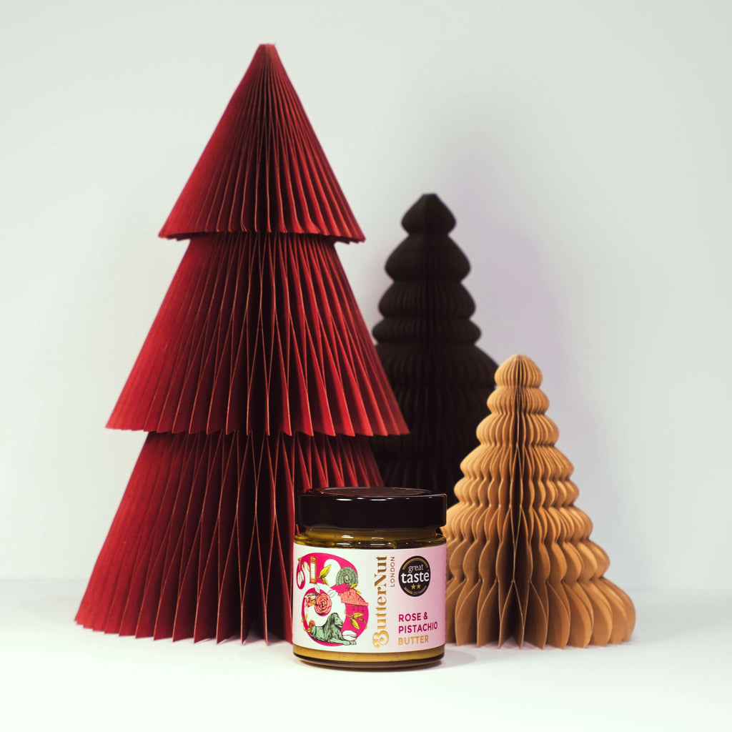 Red, green, and golden paper trees with a jar of ButterNut of London Rose & Pistachio spread on a white background.