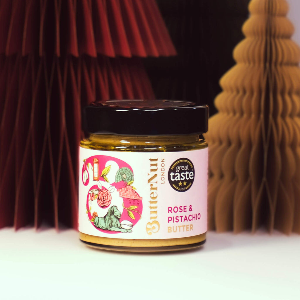 Red, green, and golden paper trees with a jar of ButterNut of London Rose & Pistachio Spread on a white background.