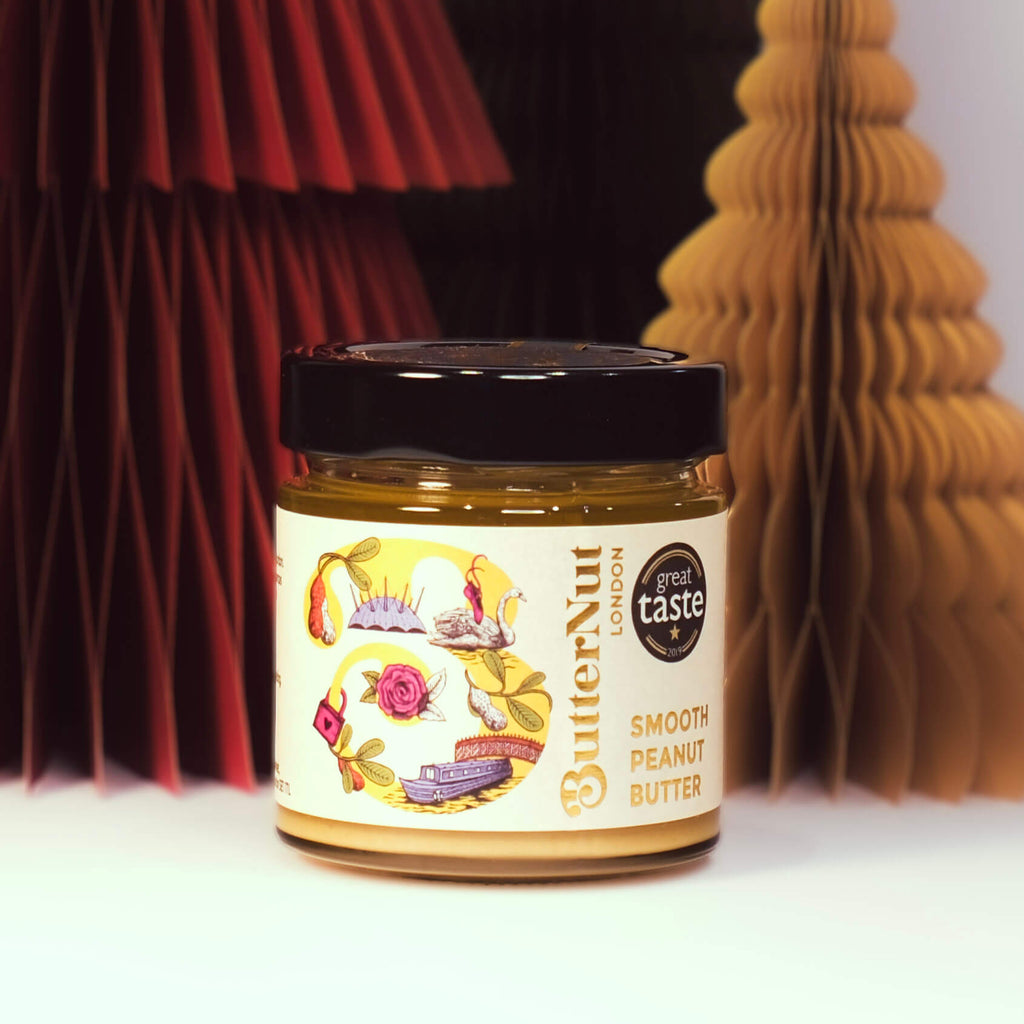 Red, green, and golden paper trees with a jar of ButterNut of London Smooth Peanut Butter on a white background.