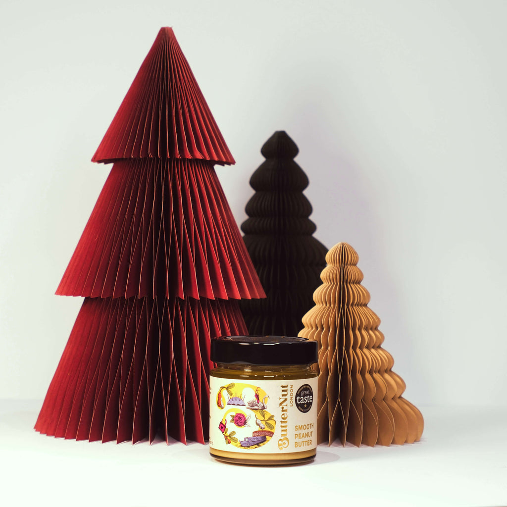 Red, green, and golden paper trees with a jar of ButterNut of London Smooth Peanut Butter on a white background.