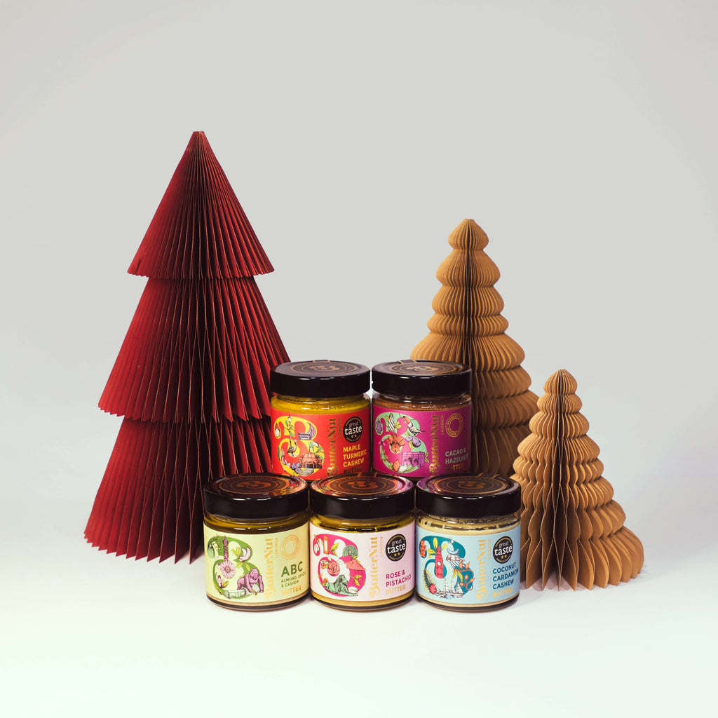 Red and golden paper trees with jars of ButterNut of London Fabulous Five Nut Butters on a white background.